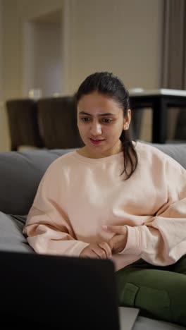 Vertical-video-of-a-happy-brunette-girl-in-light-pink-pants-communicating-via-video-conference-using-a-laptop-in-a-modern-apartment